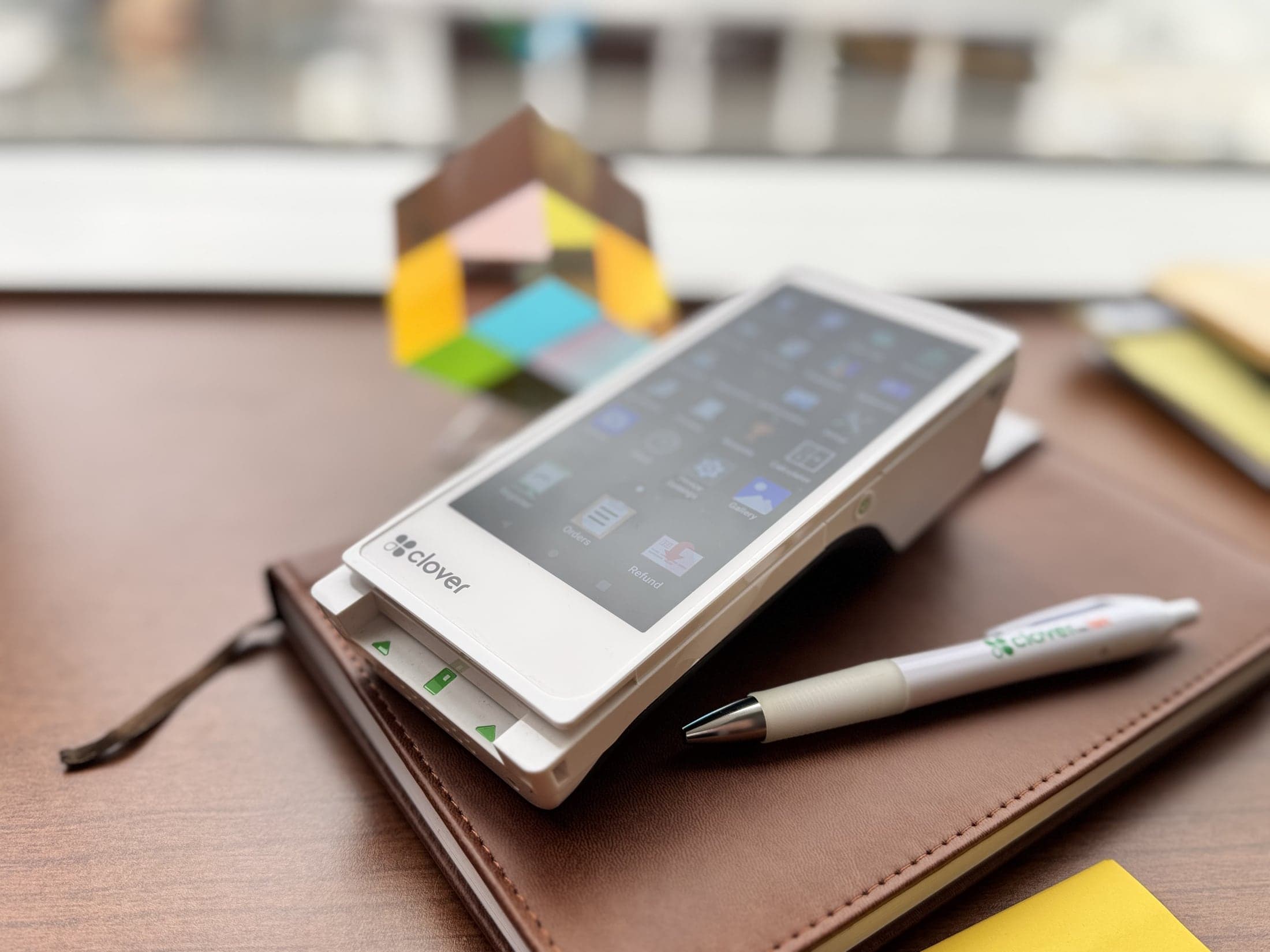 A Clover Flex payment terminal with built-in receipt printer on a desk, showing its Clover point-of-sale home screen